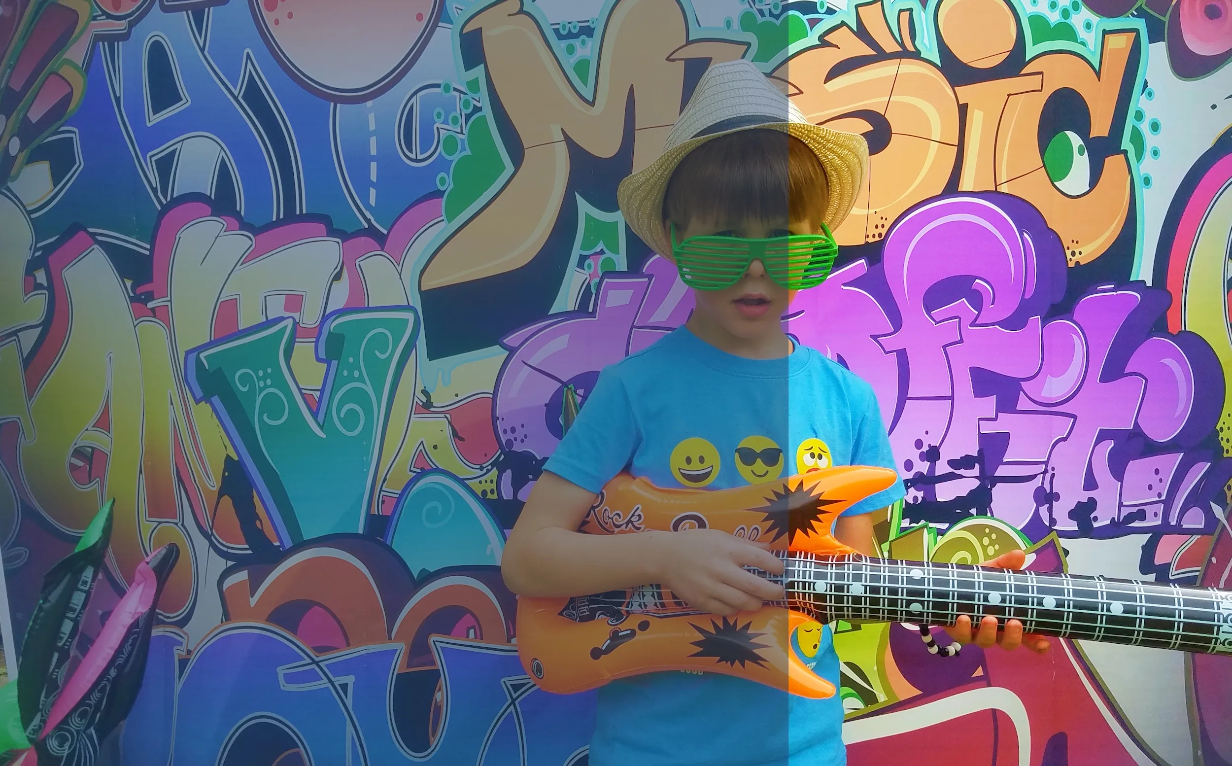 A boy in a straw hat and green shutter glasses playing an inflatable guitar in front of a colorful graffiti wall