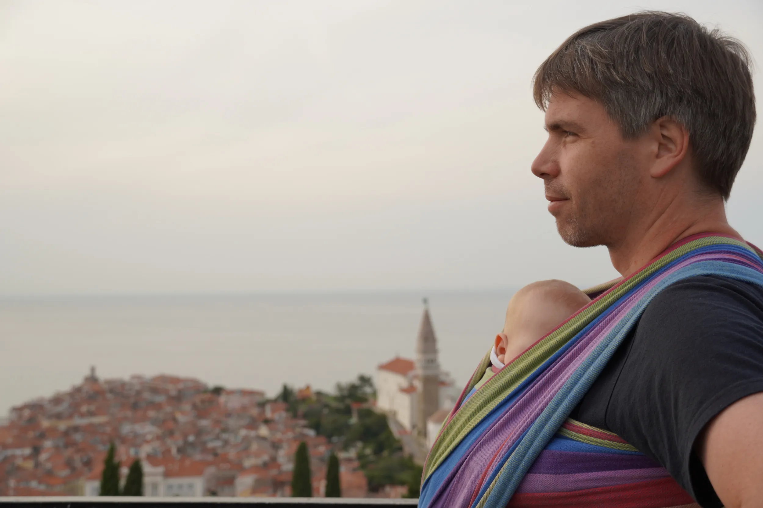 Nejc with a baby in a sling, looking out over the rooftops of Piran and the sea